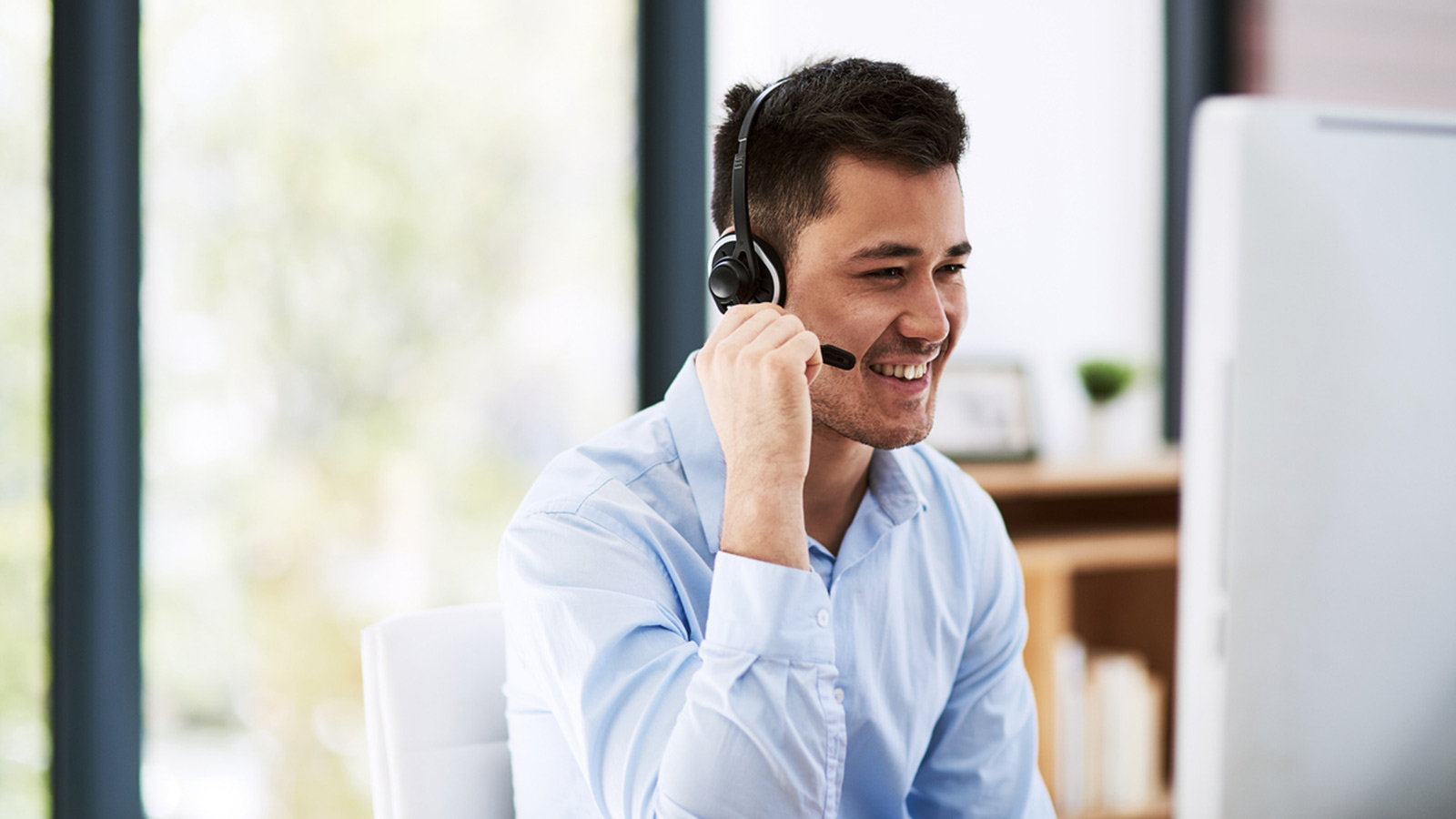 A person in business clothes smiling and wearing a headset, while sitting in front of a computer monitor. 