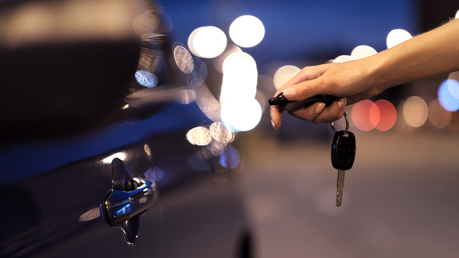 Hand holding car keys and pointing the fob towards a navy car.