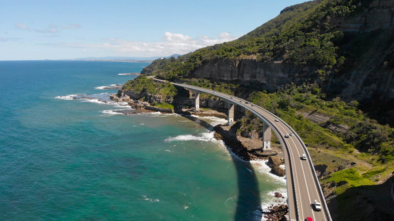 Cars driving over an ocean road on a bridge, with a greenery-covered cliff beside them.