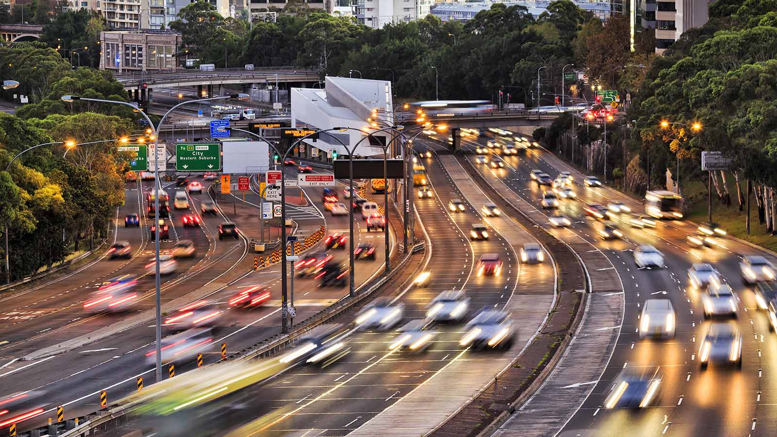 Traffic on Sydney's Warringah Freeway. There are buildings in the backgroun and green trees next to six lanes of traffic both ways.