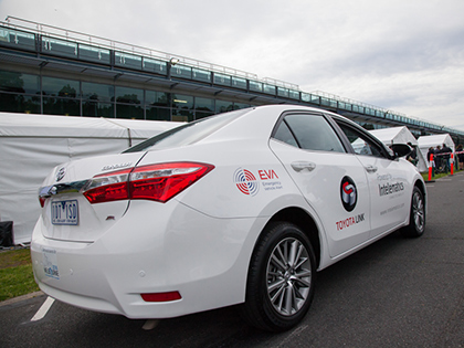 White Intelematics-branded sedan, parked on the road next to white tents set up on the grass. People are lined up in front of the tents.