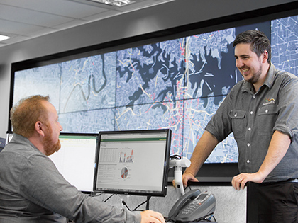 Two Intelematics staff smiling together and looking at monitors with graphs and traffic maps. One person is standing and the other is sitting at a desk.