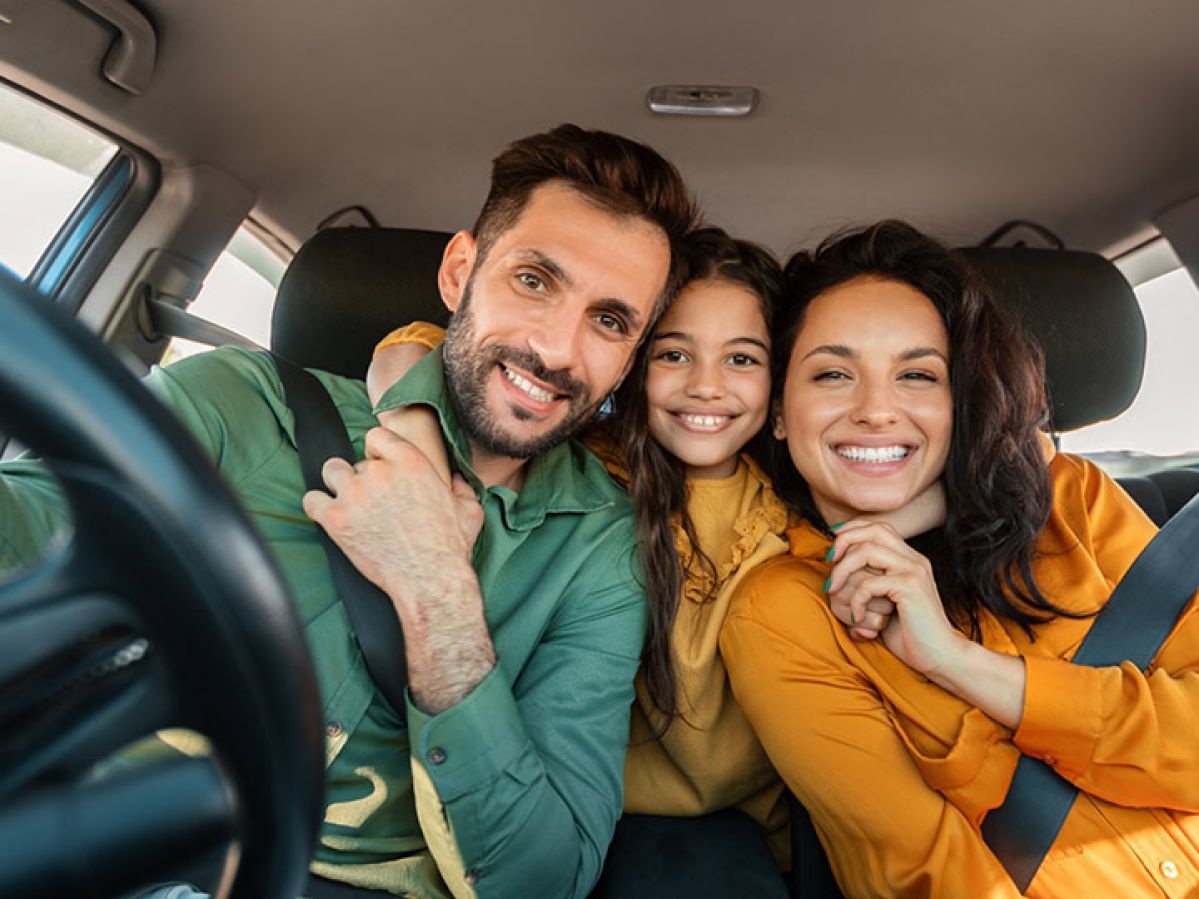 Two parents in the front seat of the car, and a child leaning from the back seat to hug them. They're all smiling together.
