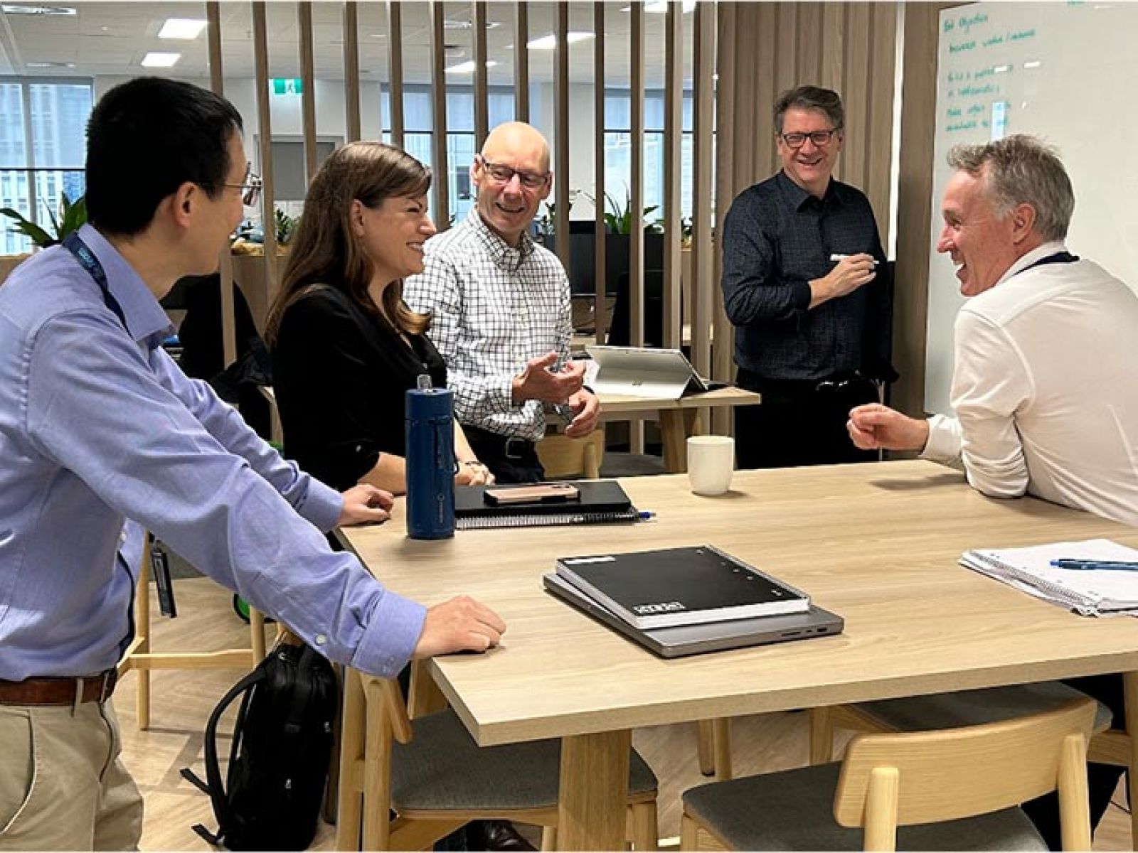 Five business people in an office, standing around a table and smiling together. One person is writing on a whiteboard.