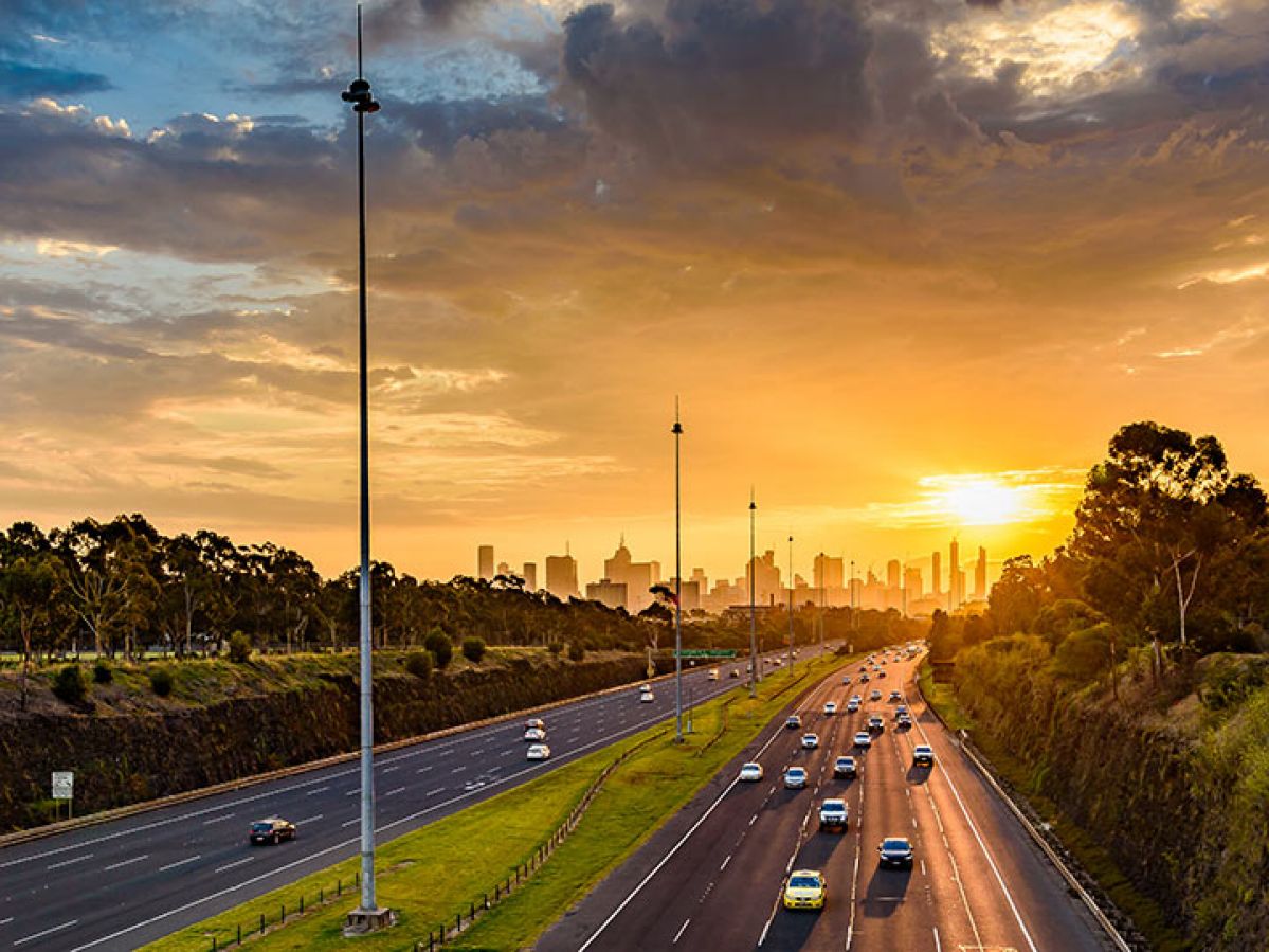 Cars on Melbourne's Eastern Freeway at sunset, with 4 lanes inbound towards the city and 5 lanes outbound. The city skyline is in the distance.