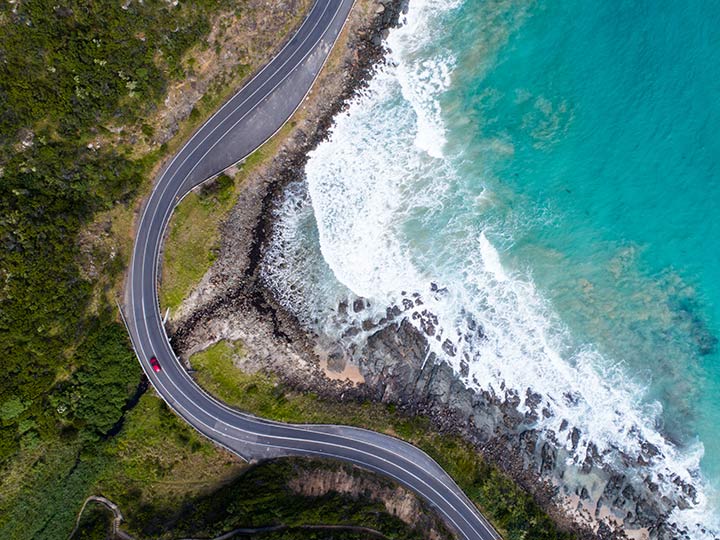 A red car driving on a road beside the ocean, with greenery on their left.