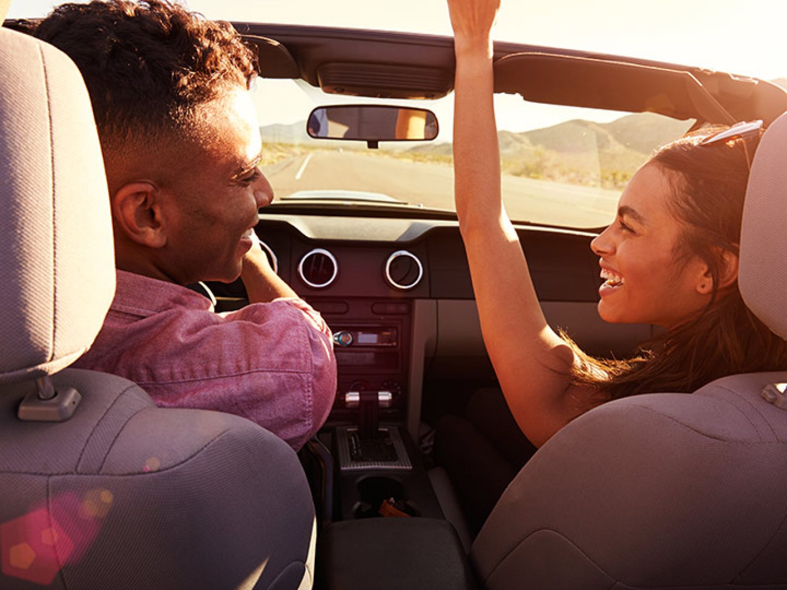 Two people in the front seat of a convertible car, smiling and looking at each other. The person in the passenger seat has one arm in the air.