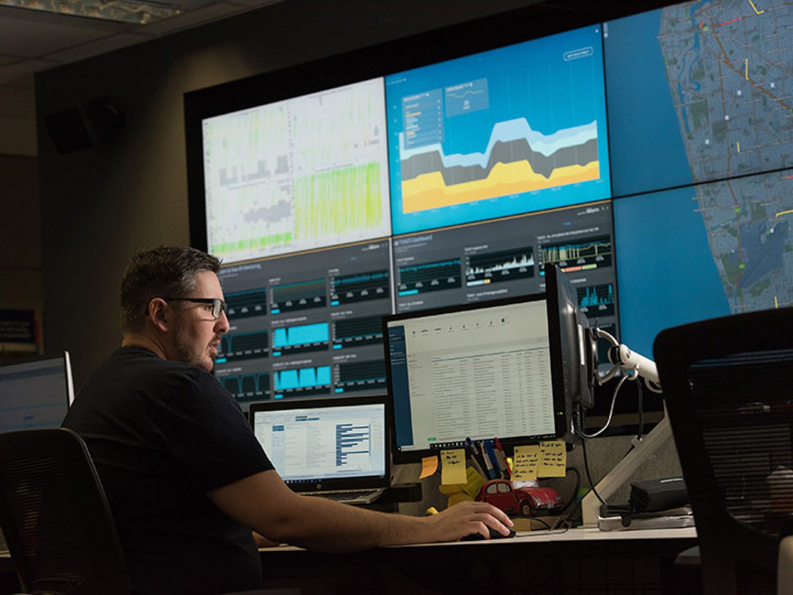 A person in business clothes sitting at their office desk, looking at monitors with graphs and traffic maps.