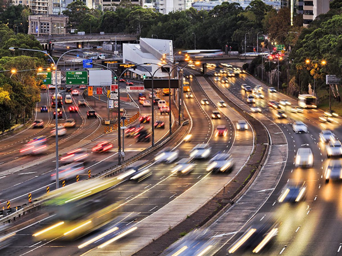 Traffic on Sydney's Warringah Freeway. There are buildings in the backgroun and green trees next to six lanes of traffic both ways.