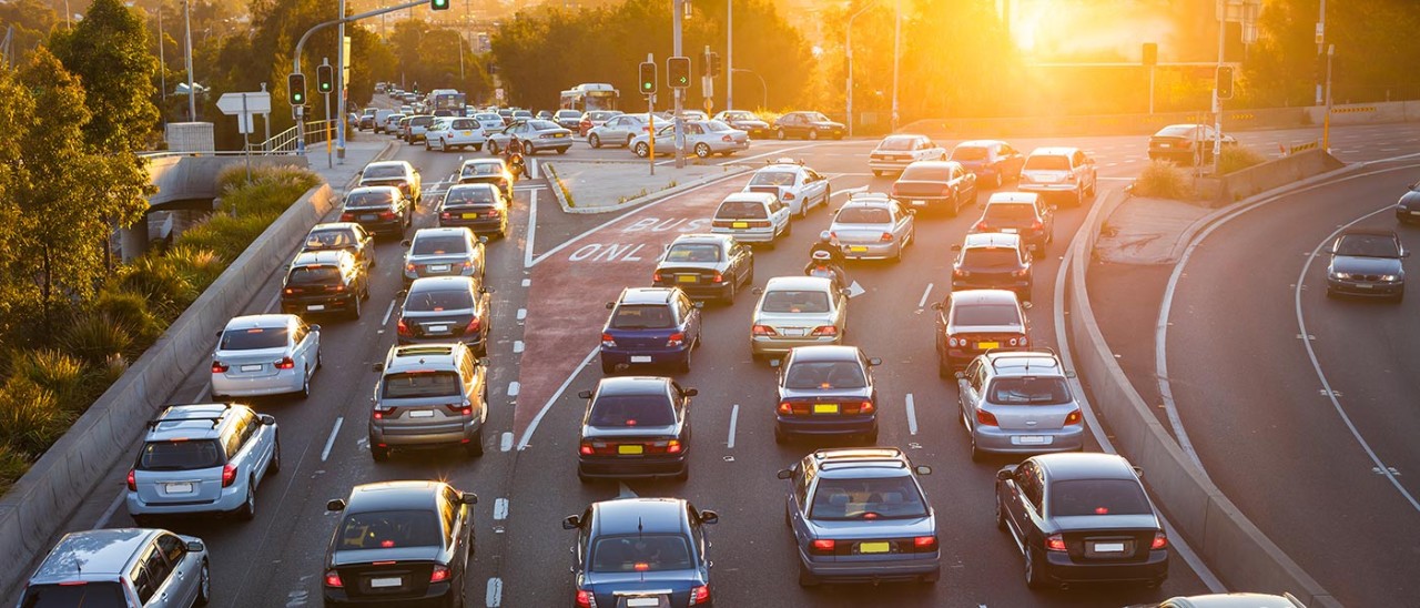 Six traffic lanes full of cars at an intersection during sunset. The first 2 lanes turn left while the other 4 lanes go straight. The far left lane going straight is a bus lane.