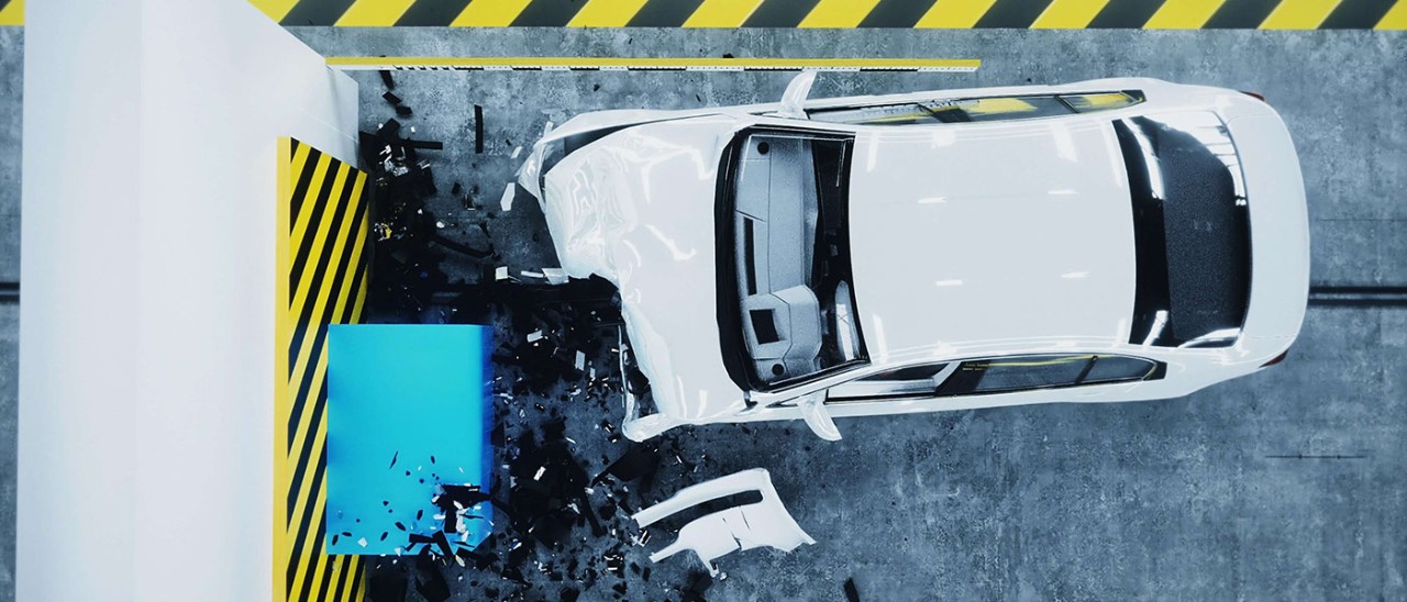 A crashed white car in a warehouse, with the front crumpled and surrounded by debris. The car is surrounded by yellow and black tape.