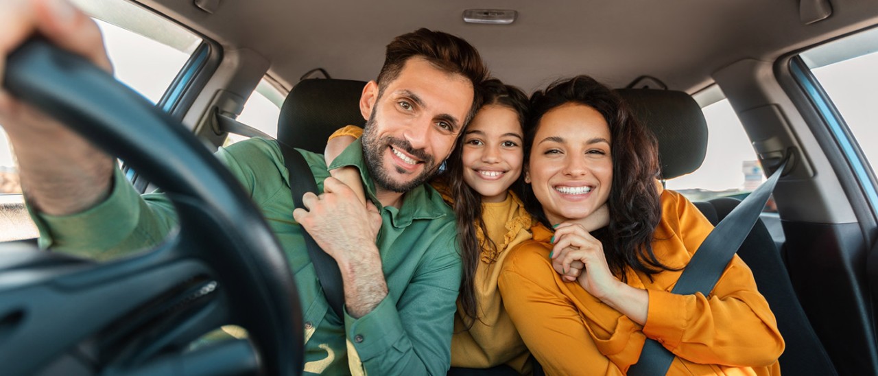 Two parents in the front seat of the car, and a child leaning from the back seat to hug them. They're all smiling together.